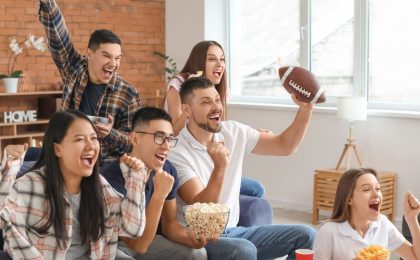 A group of people is watching a football game on TV and cheering whole eating popcorn and potato chips. We can guess they are watching football because one of the viewers is holding a football. It is a bright day, the sun is showing through the windows.