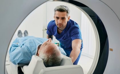 An elderly man is entering an MRI machine in a blue hospital gown with a man in darker blue scrubs attending to him. The view is from inside the MRI machine.