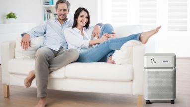 A man and woman sit together on a white couch. They are cozying up to each other, with the man’s arm around the woman and the woman reclining into the man’s body. The room is brightly lit with sunlight. The couch, room, and other decor is white.