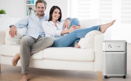 A man and woman sit together on a white couch. They are cozying up to each other, with the man’s arm around the woman and the woman reclining into the man’s body. The room is brightly lit with sunlight. The couch, room, and other decor is white.