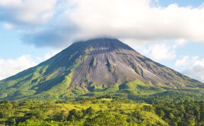 A large, towering dormant volcano covered with greenery and trees. The peak is in the clouds.