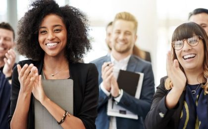 A group of people in business attire are all facing in the direction of the camera, smiling and clapping – the woman in the foreground has a tablet and a man behind her is holding a planner and pen.