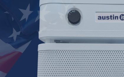 A close up of an Austin Air “it” Personal Air Purifier on the right. It is a small white appliance with a black knob and it says: “austin it” on it. There is a portion of the American flag, with red stripes and white stars on blue fabric. The flag is translucent. The background that is visible is blue.