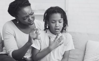 A black and white photo showing a mother and daughter sitting on a couch during an asthma attack. The child is holding her chest and her mother is rubbing her back with one hand while holding an inhaler at-the-ready in the other hand.