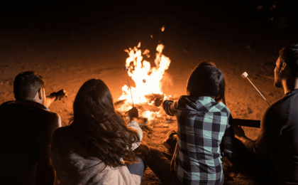 A photo of a nighttime bonfire on the beach. Four people, who appear to be two men and two women, are sitting with their backs to the camera. They are roasting marshmallows.